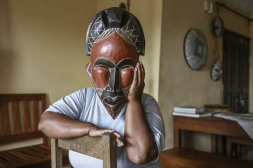 Une femme assise tient un masque de bois sculpté devant son visage